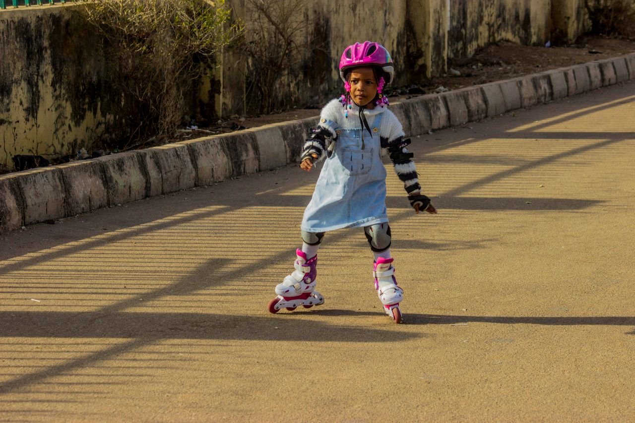 Mastering the First Impression: Your intriguing post title goes here A young girl in protective gear roller skating on a sunny day, wearing a pink helmet and blue dress.